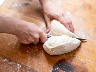 Female hands cut with a knife a kneaded dough on a wooden table. The process of preparing fresh elastic baking dough. Table and hands in flour. Natural home cooking.	