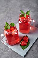 Homemade strawberry tea with mint in mason jar on gray concrete table background, copy space. Cold summer berry drink in sparkling glasses. Fresh vitamin beverage