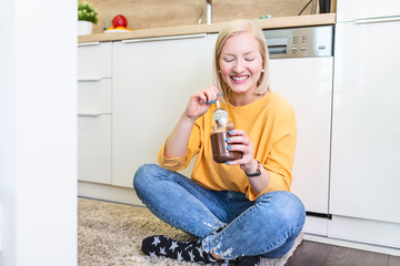 Young woman eating chocolate from a jar while sitting on the wooden kitchen floor. Cute albino girl indulging cheeky face eating chocolate spread from jar using spoon savoring every mouthful