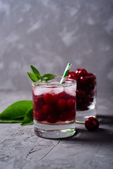 Fresh cherry lemonade with ice, mint and paper straw in sparkling glasses on gray table background, copy space. Cold summer drink. Berry cocktail