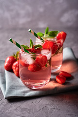 Fresh strawberry lemonade with ice, mint and paper straw in sparkling glasses on gray table background, copy space. Cold summer drink. Berry cocktail