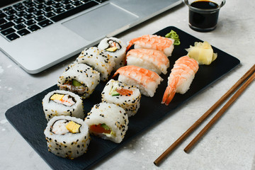 food delivery set of sushi and rolls with salmon and shrimp on a gray table next to a laptop. lunch at the computer. the concept of smart office. Top view of sushi, uramaki, hosomaki and nigiri. 