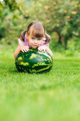 Little toddler girl squatting by a large watermelon on the lawn in the summer garden