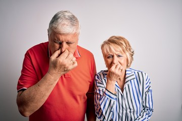 Senior beautiful couple standing together over isolated white background smelling something stinky and disgusting, intolerable smell, holding breath with fingers on nose. Bad smell