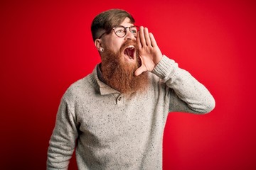 Handsome Irish redhead man with beard wearing casual sweater and glasses over red background shouting and screaming loud to side with hand on mouth. Communication concept.