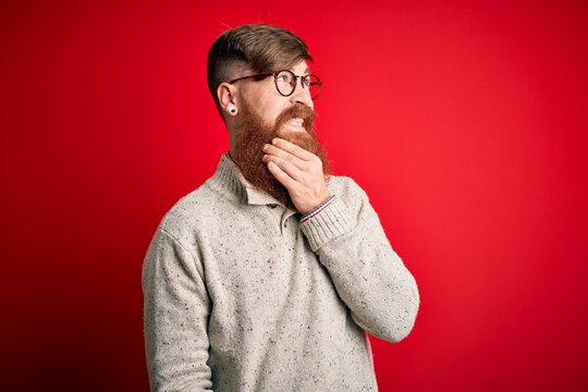Handsome Irish redhead man with beard wearing casual sweater and glasses over red background Thinking worried about a question, concerned and nervous with hand on chin