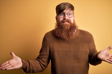 Handsome Irish redhead man with beard wearing glasses and winter sweater over yellow background...