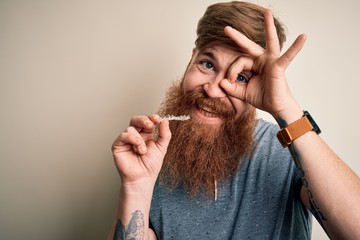 Irish redhead man with beard holding dental invisible aligner for tooth correction with happy face smiling doing ok sign with hand on eye looking through fingers