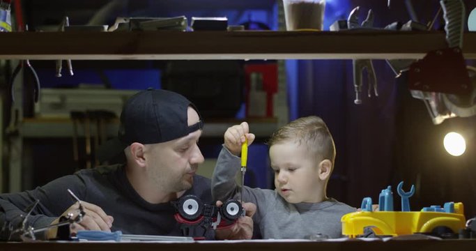 Father and Son Fixing Together a Toy Tractor with Screwdriver in Garage and Little Boy is Learning to Work with Tools Shot on Red Camera
