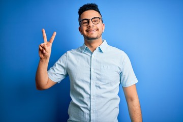 Young handsome man wearing casual summer shirt and glasses over isolated blue background smiling with happy face winking at the camera doing victory sign. Number two.