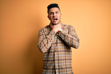 Young handsome man wearing casual shirt standing over isolated yellow background shouting suffocate because painful strangle. Health problem. Asphyxiate and suicide concept.