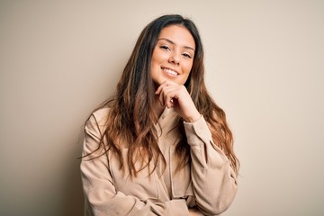 Young beautiful brunette woman wearing casual shirt standing over white background looking confident at the camera with smile with crossed arms and hand raised on chin. Thinking positive.