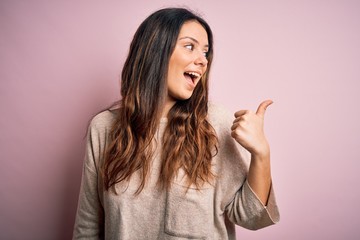 Young beautiful brunette woman wearing casual sweater standing over pink background smiling with happy face looking and pointing to the side with thumb up.