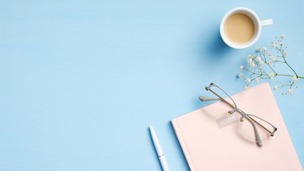 Modern office desk with pink notebook, pen, glasses, cup of coffee, flowers on blue background. Minimal style feminine workspace, business and education concept. Flat lay, top view