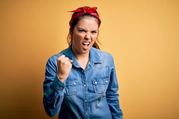 Young beautiful brunette woman wearing casual denim shirt and hair handkerchief angry and mad raising fist frustrated and furious while shouting with anger. Rage and aggressive concept.