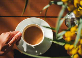 female hand takes a white cup of coffee from the table, on the table is a bouquet with mimosa, view and shot from above