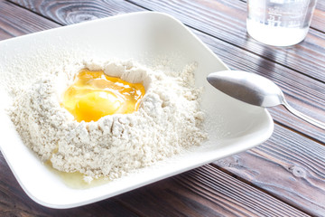 Broken egg in flour in white plate, spoon on plate. Sun's rays on table.