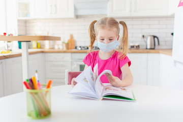 Little blonde girl with two ponytales reading a book at home in the kitchen wearing medical pritective face mask to protect herself from the virus