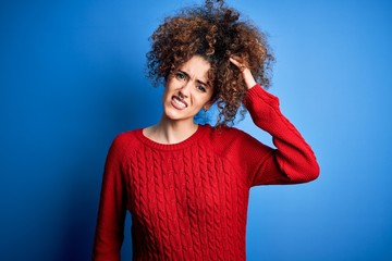 Fototapeta na wymiar Young beautiful woman with curly hair and piercing wearing casual red sweater confuse and wonder about question. Uncertain with doubt, thinking with hand on head. Pensive concept.