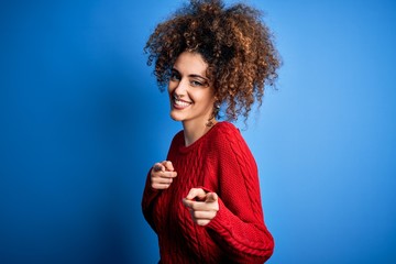 Young beautiful woman with curly hair and piercing wearing casual red sweater pointing fingers to camera with happy and funny face. Good energy and vibes.