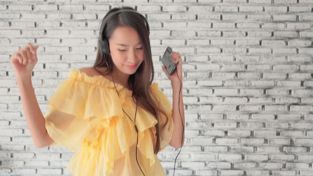 Young attractive Asian woman in yellow casual shirt and jeans against white brick wall delightfully listening to music on headphones cable connected to mobile phone. 