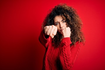 Young beautiful woman with curly hair and piercing wearing casual red sweater Punching fist to fight, aggressive and angry attack, threat and violence