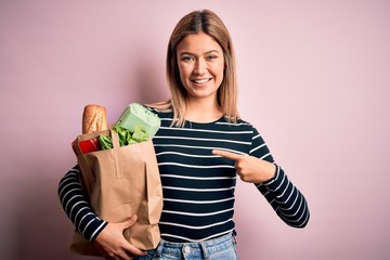 Young beautiful woman holding paper bag with purchase over isolated pink background with surprise face pointing finger to himself