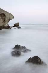Long exposure photograph, with a rock formation in the foreground and a cliff in the background, taken on the beach of La Media Luna in the Cabo de Gata Natural Park, Almeria, Andalusia.