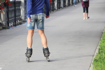 Fototapeta na wymiar A man in shorts without safety equipment skating with rollerblades on a asphalt road alley in Park at summer day, waist-high rear view close up, city vacation activity outdoors 