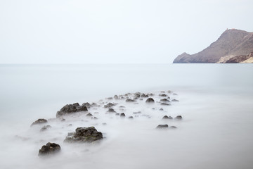 Long exposure photograph, with a rock formation in the foreground and a cliff in the background, taken on the beach of Monsul in the Cabo de Gata Natural Park, Almeria, Andalusia.