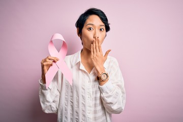 Young beautiful asian girl holding pink cancer ribbon symbol over isolated background cover mouth with hand shocked with shame for mistake, expression of fear, scared in silence, secret concept