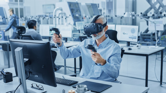 Industrial Design Engineer Wearing Virtual Reality Headset and Holding Controllers, Uses VR technology for Industrial Design, Development using Computer. Modern Factory Office, Workshop with Machinery