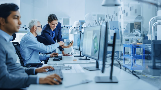 Inside Factory Office: Female Project Supervisor Talks to Male Senior Industrial Engineer who Works on Computer, Talk. In Workshop: Professional Workers Use High-Tech Industry CNC Machinery