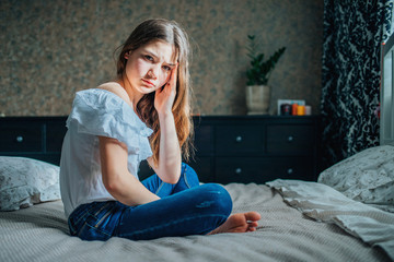 Obraz premium a sad dark-haired girl in a white blouse and blue jeans is sitting on the bed in her bedroom. She has a headache. horizontal shot, selective focus.