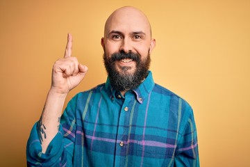 Naklejka premium Handsome bald man with beard wearing casual shirt standing over isolated yellow background pointing finger up with successful idea. Exited and happy. Number one.