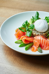 Tasty and healthy continental breakfast - quinoa with benedict eggs, spinach, avocado, salted salmon and asparagus in white bowl. Wooden background