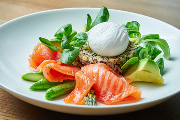 Tasty and healthy continental breakfast - quinoa with benedict eggs, spinach, avocado, salted salmon and asparagus in white bowl. Wooden background
