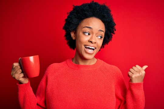 African American afro woman with curly hair drinking cup of coffee over red background pointing and showing with thumb up to the side with happy face smiling