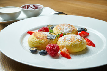Classic Ukrainian syrniki with berries, jam and sour cream in a white plate on a wooden background. Appetizing breakfast dessert