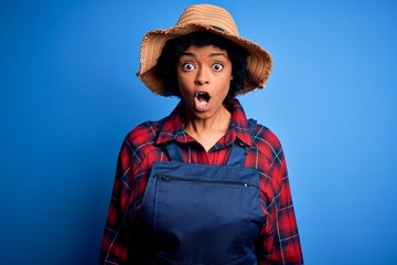 Young African American afro farmer woman with curly hair wearing apron and hat afraid and shocked with surprise expression, fear and excited face.