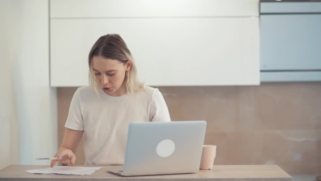 A young girl is concentrated in her laptop and compares information with what is written on a piece of paper. Bright spacious room.