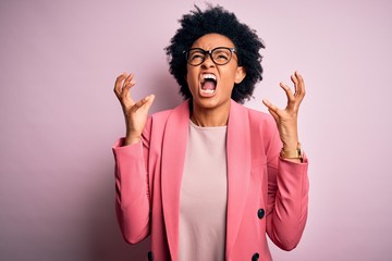 Young beautiful African American afro businesswoman with curly hair wearing pink jacket crazy and mad shouting and yelling with aggressive expression and arms raised. Frustration concept.