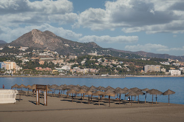 Malaga Beach La Malagueta panorama with straw beach umbrellas in the sand. Mountains and hills in the sea background in Costa del Sol, Spain.