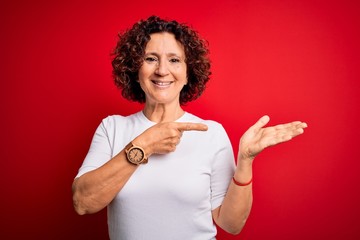 Middle age beautiful curly hair woman wearing casual t-shirt over isolated red background amazed and smiling to the camera while presenting with hand and pointing with finger.