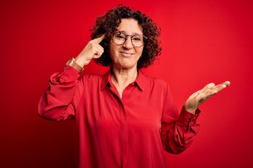 Middle age beautiful curly hair woman wearing casual shirt and glasses over red background confused and annoyed with open palm showing copy space and pointing finger to forehead. Think about it.