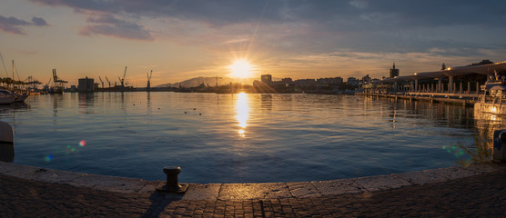 Sunset in Malaga Port wide panorama from Quay 1 in Spain with sun reflections in the sea water and cranes.