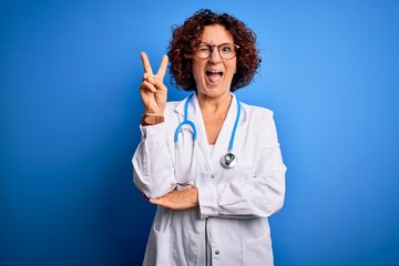 Middle age curly hair doctor woman wearing coat and stethoscope over blue background smiling with happy face winking at the camera doing victory sign. Number two.