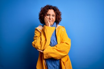 Middle age curly hair woman wearing rain coat standing over isolated blue background thinking looking tired and bored with depression problems with crossed arms.