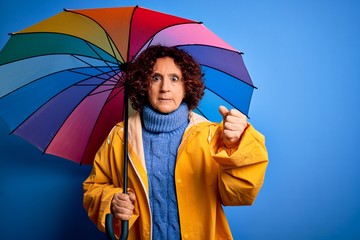 Middle age curly hair woman wearing rain coat holding colorful umbrella over blue background annoyed and frustrated shouting with anger, crazy and yelling with raised hand, anger concept