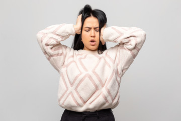 Portrait of a beautiful brunette in a warm white winter sweater. The young woman has a headache, on a gray background. The girl holds her hands behind her head